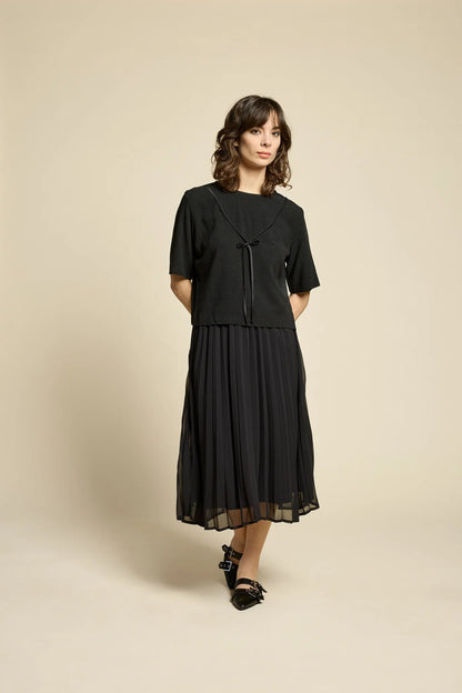A woman wearing the Flowers Skirt by Cokluch in Black, a midi-length pleated skirt with a sheer overlayer and an elastic waist. She is wearing it with a black top  and standing in front of a beige background. 