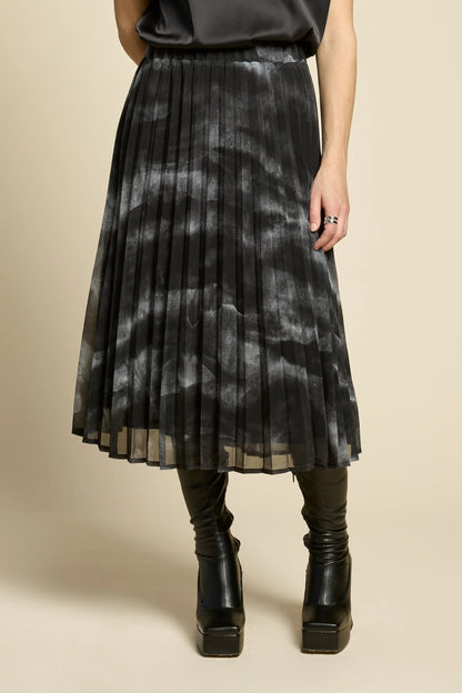 Waist down view of a woman wearing the Flowers Skirt by Cokluch in Mist Black, a midi-length pleated skirt with a sheer over-layer and an elastic waist. She is wearing it with a black top and standing in front of a beige background. 
