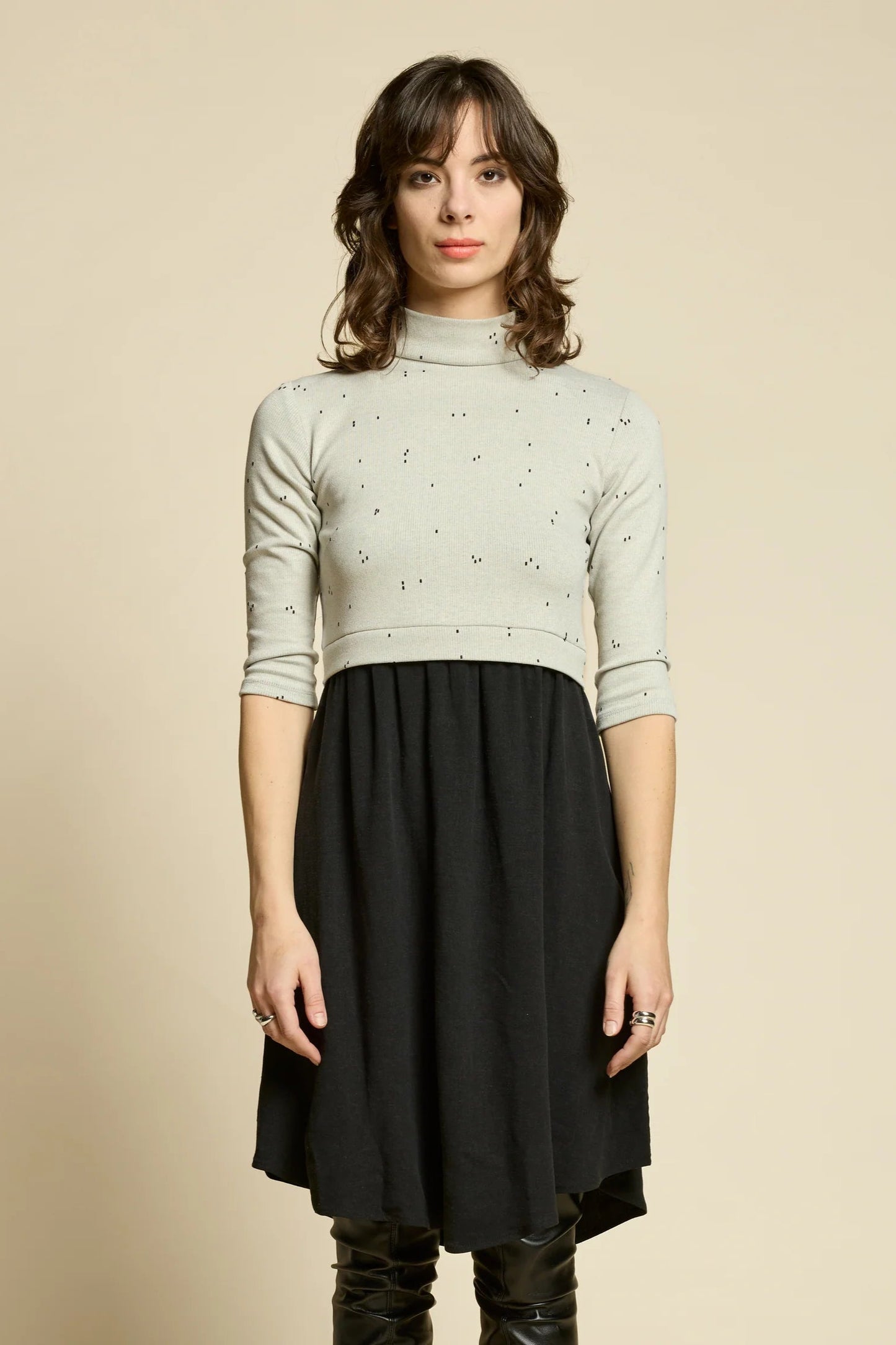 A woman wearing the Crystal Dress by Cokluch, featuring a knit top half in Natural with a flowing black skirt that falls to the knee. It has a high neck, 3/4 sleeves, and an opening at the back. She is standing in front of a beige background. 