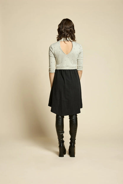 Back view of  woman wearing the Crystal Dress by Cokluch, featuring a knit top half in Natural with a flowing black skirt that falls to the knee. It has a high neck, 3/4 sleeves, and an opening at the back. She is standing in front of a beige background. 