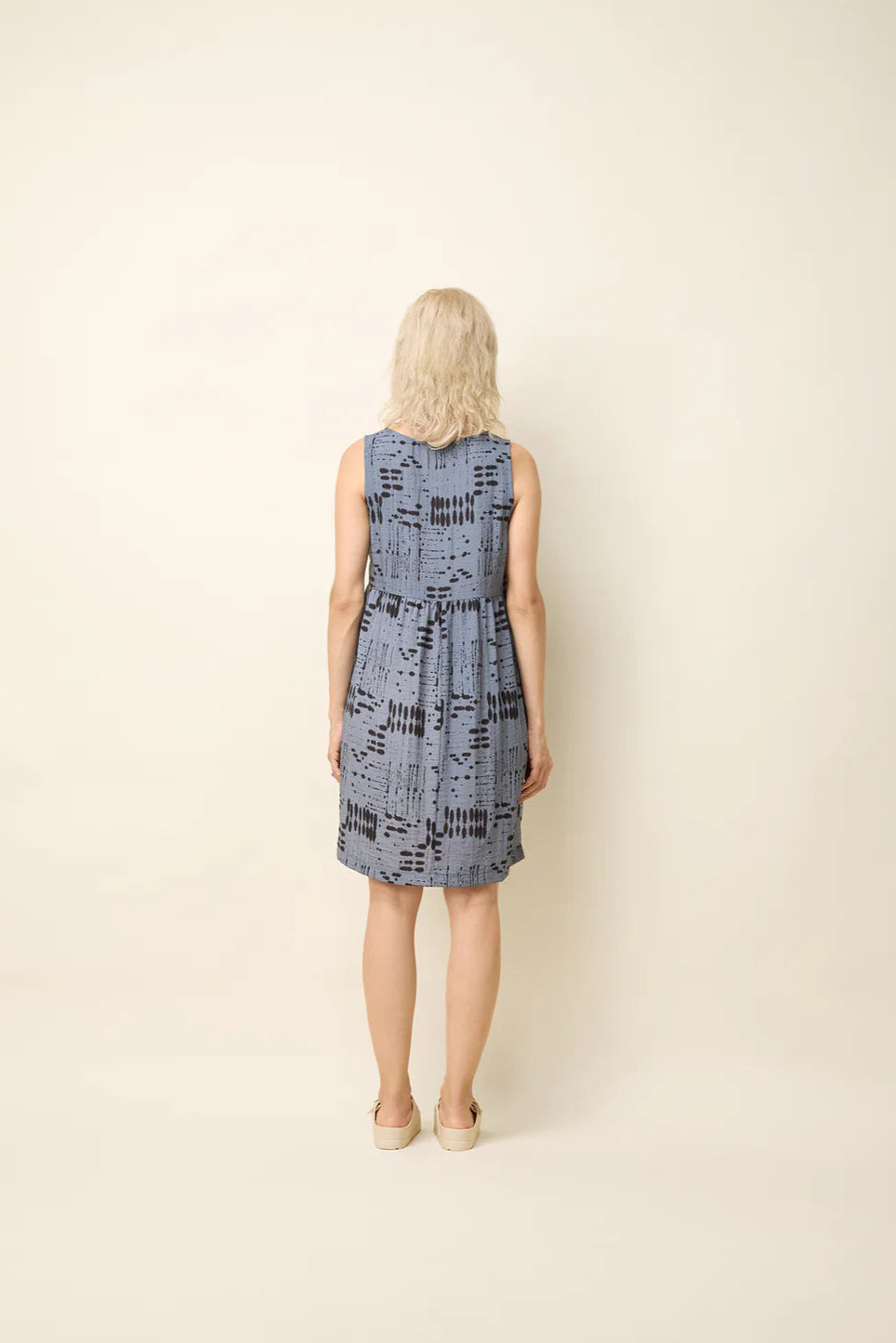 Back view of A woman wearing the Esi Dress by Cokluch in Blue Ink, a sleeveless above-the-knee dress with a round neck, a triangle cutout below the neckline, a empire waist, and a loose fit. She's standing in front of a beige background. 