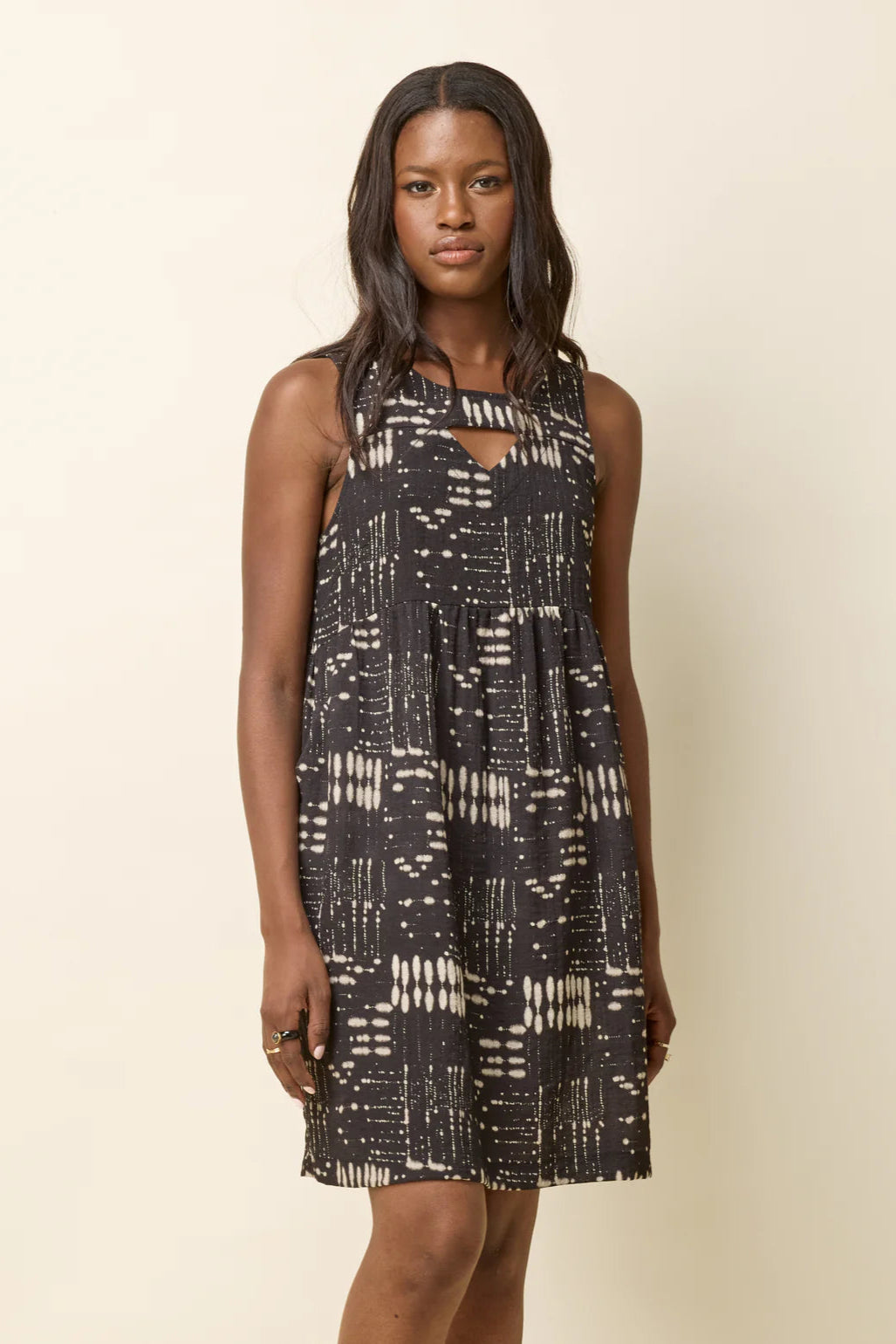 A woman wearing the Esi Dress by Cokluch in Black Ink, a sleeveless above-the-knee dress with a round neck, a triangle cutout below the neckline, a empire waist, and a loose fit. She's standing in front of a beige background. 