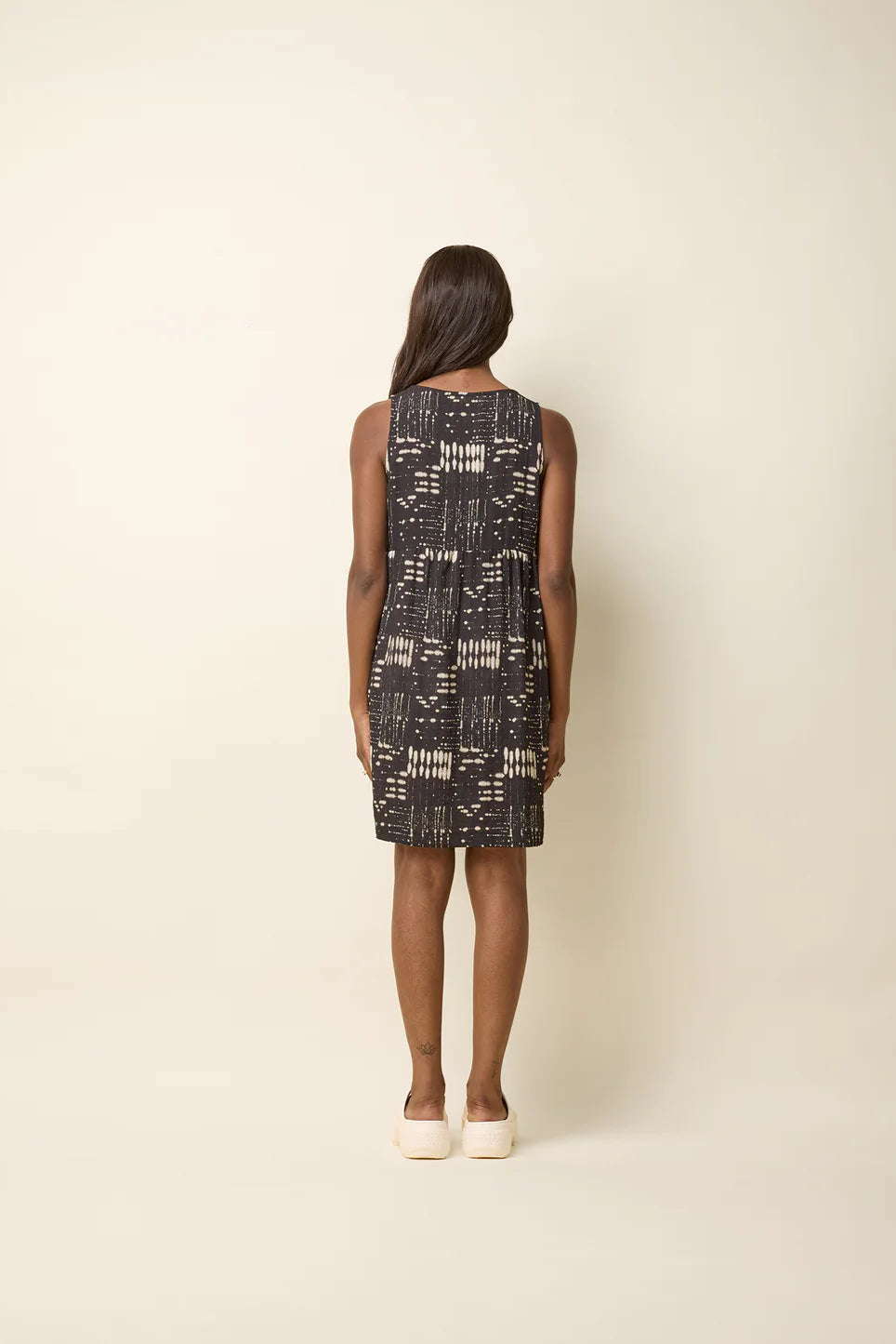 Back view of a woman wearing the Esi Dress by Cokluch in Black Ink, a sleeveless above-the-knee dress with a round neck, a triangle cutout below the neckline, a empire waist, and a loose fit. She's standing in front of a beige background. 