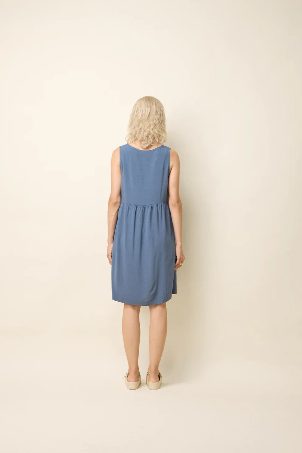 Back view of a woman wearing the Esi Dress by Cokluch in Ocean, a sleeveless above-the-knee dress with a round neck, a triangle cutout below the neckline, a empire waist, and a loose fit. She's standing in front of a beige background. 