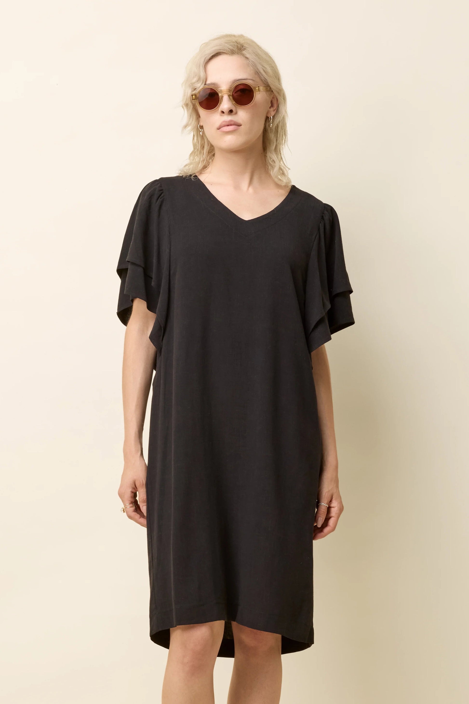 A woman wearing the Paramida Dress by Cokluch in Black, featuring a v-neck, a double ruffle on the short sleeves, and a loose fit that falls above the knee. She is standing in front of a beige background. 