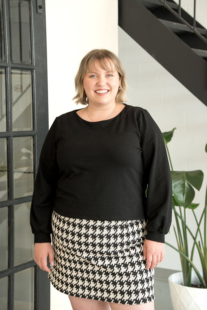 A woman wearing the Cortado Skirt by Cherry Bobin in Houndstooth, a short fitted skirt with an elastic waistband and invisible side zipper. She is wearing it with a black top and standing in front of a plant. 