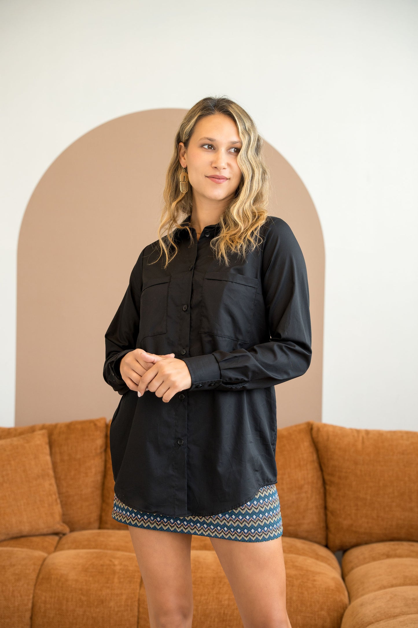A woman wearing the Cortado Skirt by Cherry Bobin in Jaquard Zigzag, a short fitted skirt with an elastic waistband and invisible side zipper. She is wearing it with a black top and standing in front of a couch.
