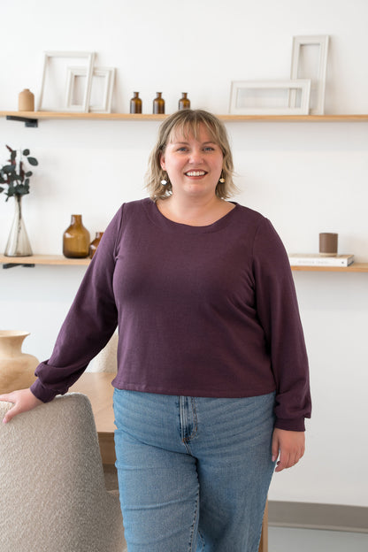 A woman wearing the Cosmopolitain Sweater by Cherry Bobin in Mauve, with a rounded neck and long sleeves with a puff detail. Shbe is wearing it with jeans and standing in a room with shelves. 