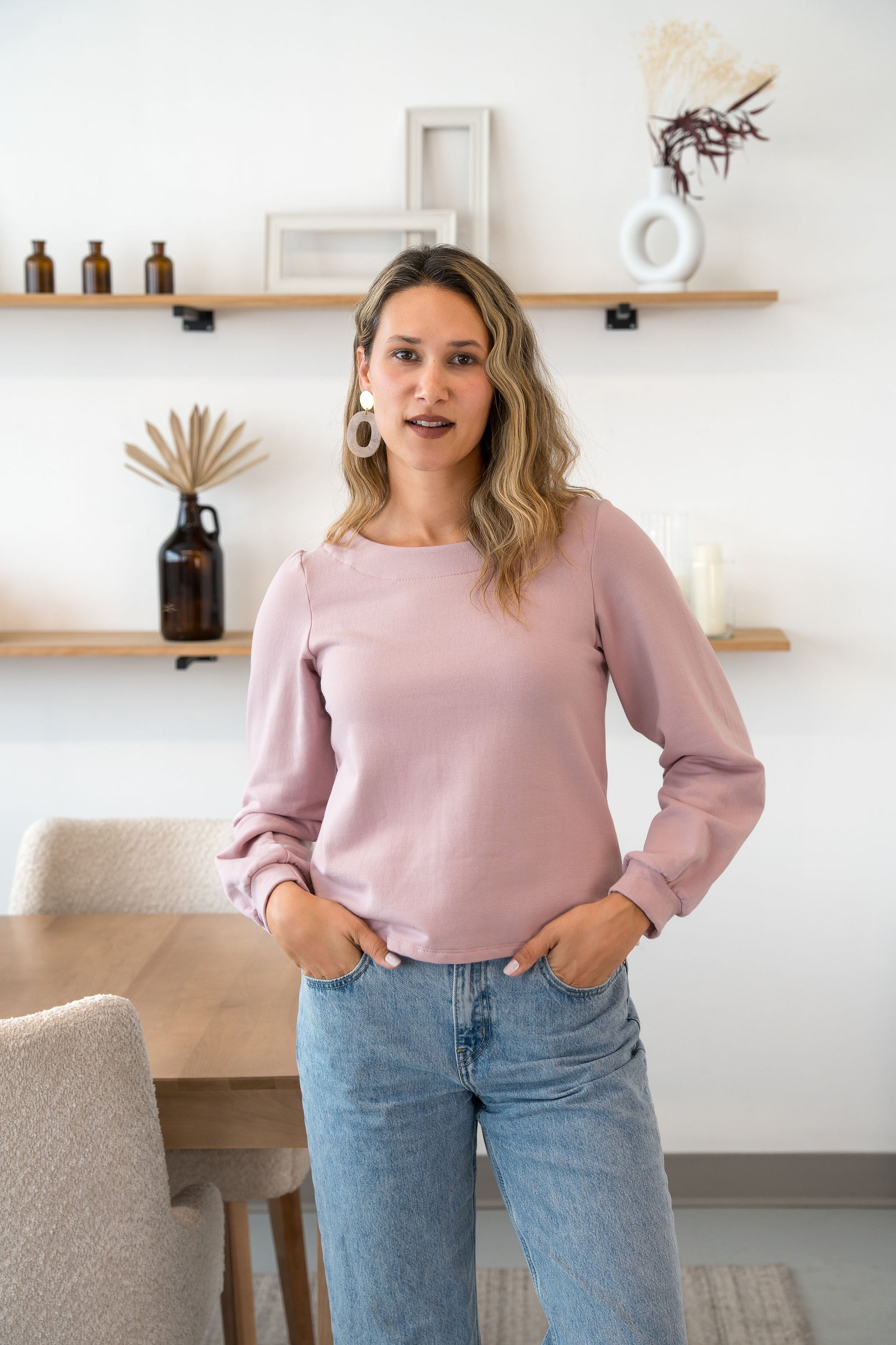 A woman wearing the Cosmopolitain Sweater by Cherry Bobin in Rose, with a rounded neck and long sleeves with a puff detail. Shbe is wearing it with jeans and standing in a room with shelves. 