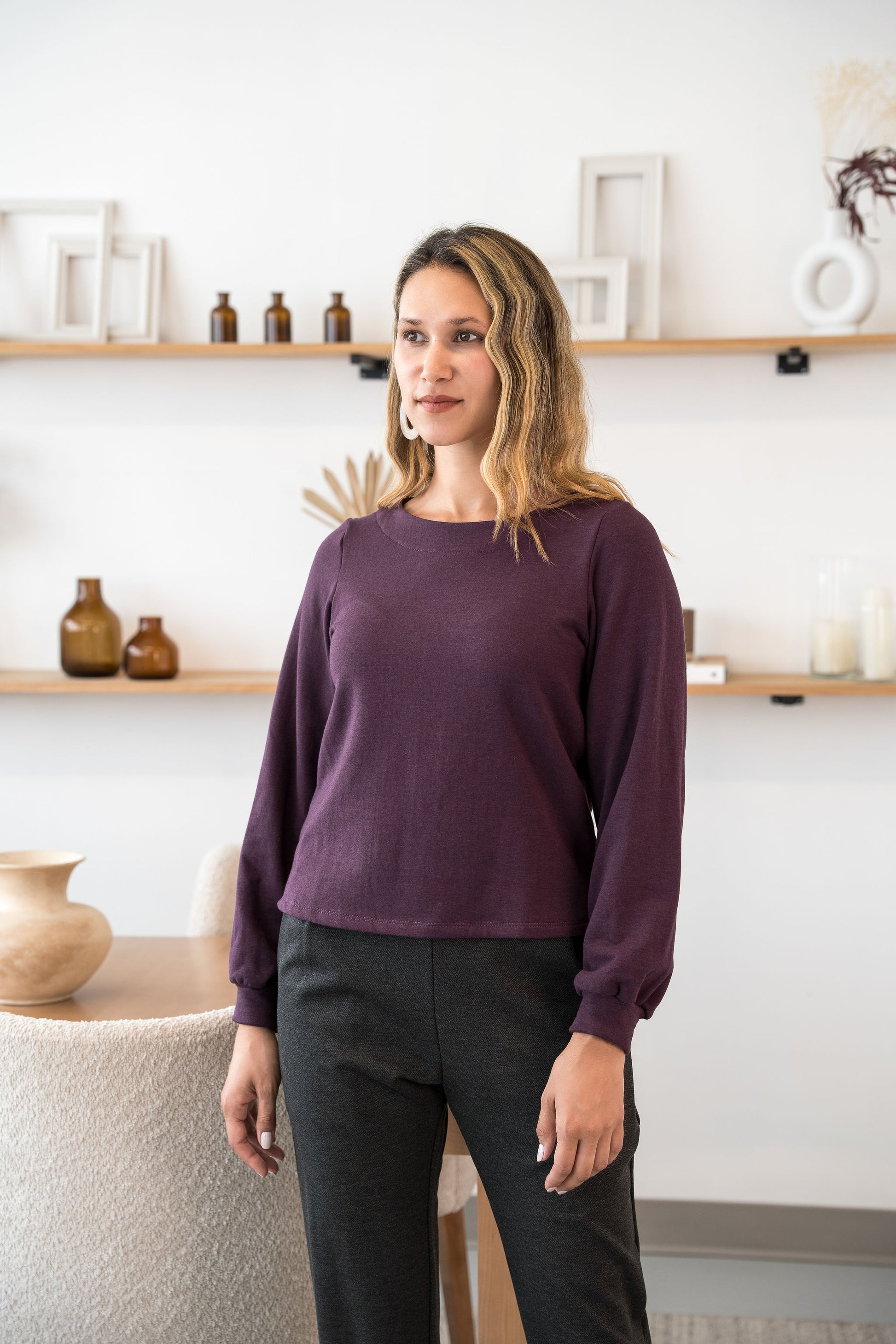 A woman wearing the Cosmopolitain Sweater by Cherry Bobin in Mauve, with a rounded neck and long sleeves with a puff detail. She is wearing it with black pants and standing in a room with shelves. 