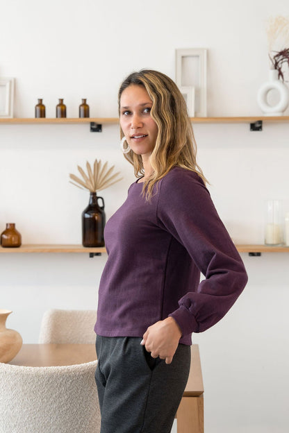 A woman wearing the Cosmopolitain Sweater by Cherry Bobin in Mauve, with a rounded neck and long sleeves with a puff detail. She is wearing it with black pants and standing in a room with shelves. 