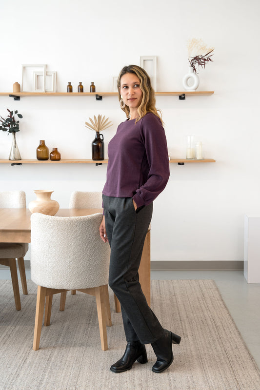 A woman wearing the Manhattan Pants by Cherry Bobin in Grey, straight leg Ponte di Roma pants with a pull on waist. She is wearing them with the Cosmopolitain Sweater  and standing in a room with shelves. 