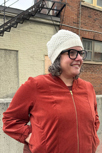 A woman wearing the Beret by Charlevoix Pure Laine in Natural with the Redrawn Zip Jacket by Eve Lavoie in Terracotta Corduroy. She is standing outside. 