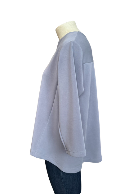 A side view of the Vanessa Top by Solomia in Blue is shown on a mannequin against a white background. It has a notched neckline, 3/4 sleeves, a rounded hi-low hemline, and a vertical seam down the front. 