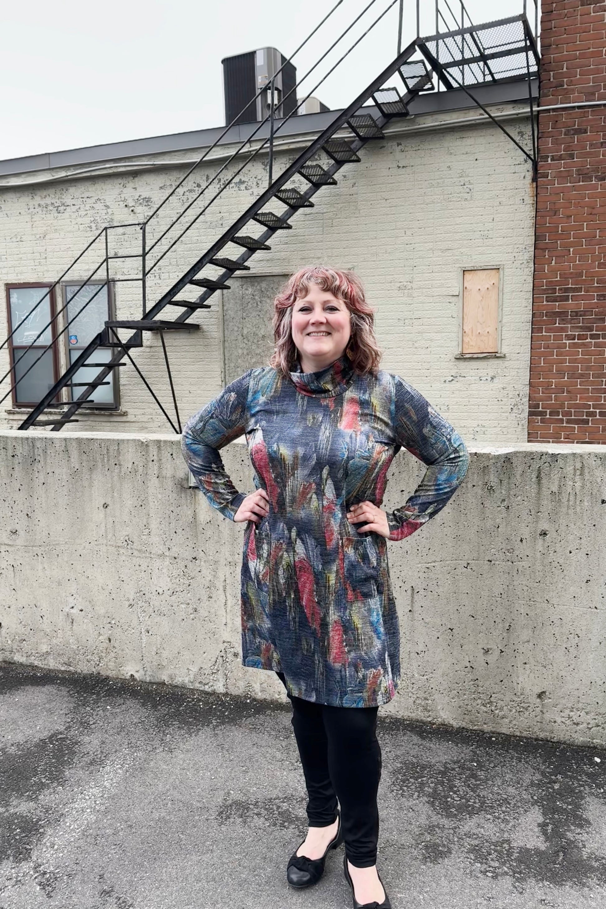 A woman wearing the Dakota Dress by Pure Essence, featuring a cowl neck, long sleeves, patch pockets, a knee-length hem, and a subtly sparkly Multicolour Print. She's wearing black leggings and standing outside in front of a brick building. 