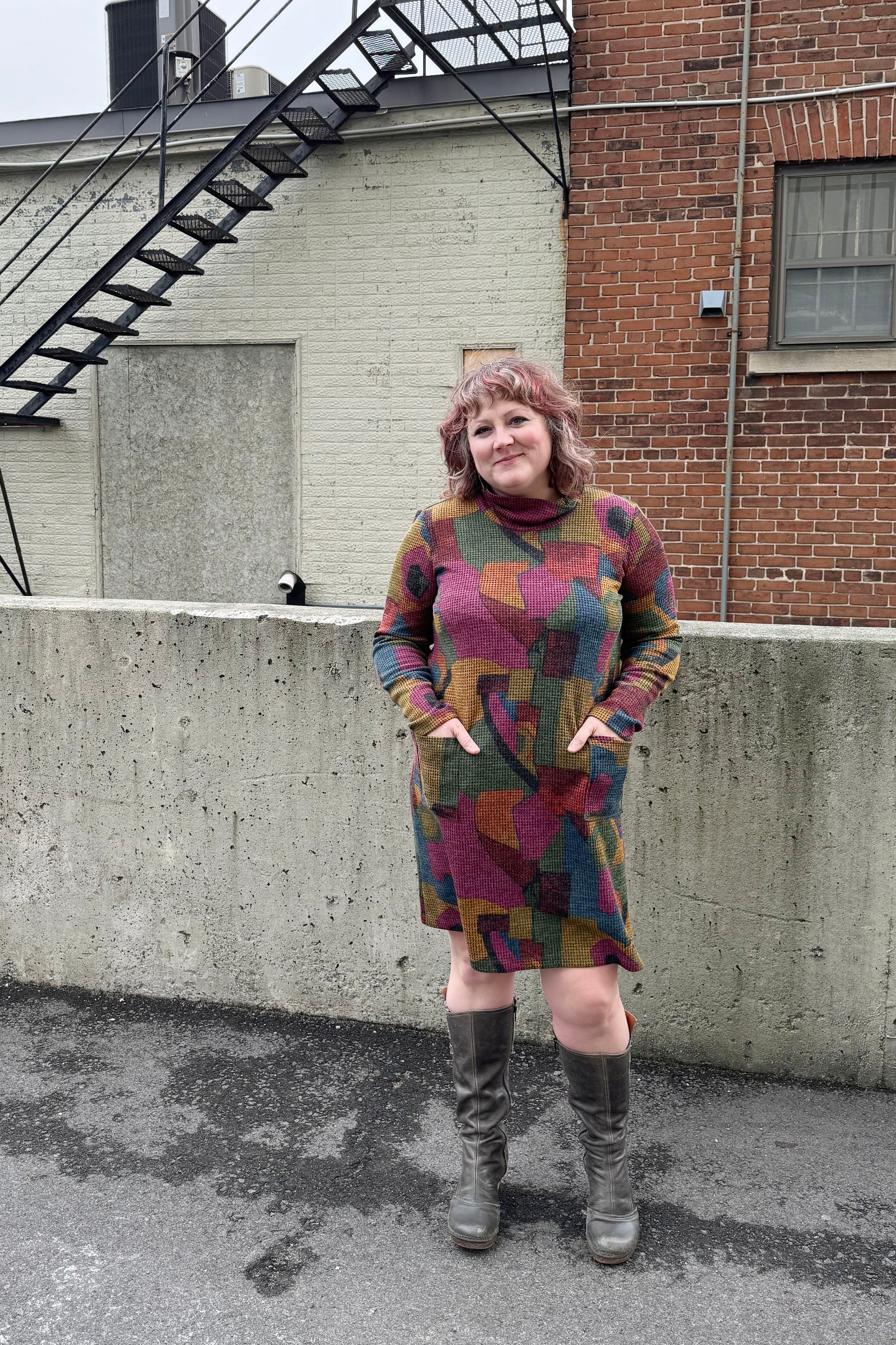 A woman wearing the Damon Dress by Pure Essence in Berry/Mustard, featuring a cowl neck, long sleeves, patch pockets, and a knee length hem. She's standing outside in front of a brick building. 