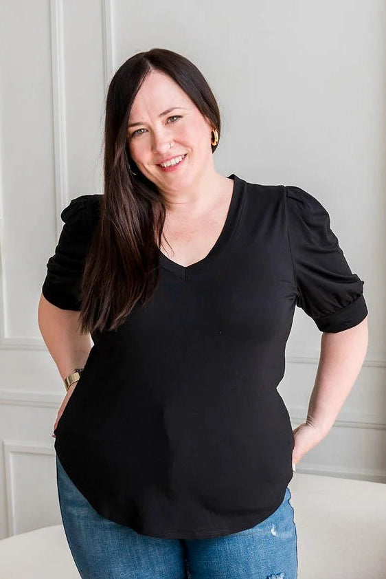 A woman wearing the Darling Tee by Dotty in Black, featuring a v-neck, pleated shoulders, and elbow length sleeves, with jeans. She is standing n front of a white sofa in a room with white walls 