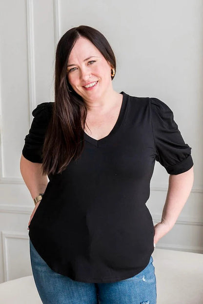 A woman wearing the Darling Tee by Dotty in Black, featuring a v-neck, pleated shoulders, and elbow length sleeves, with jeans. She is standing n front of a white sofa in a room with white walls 