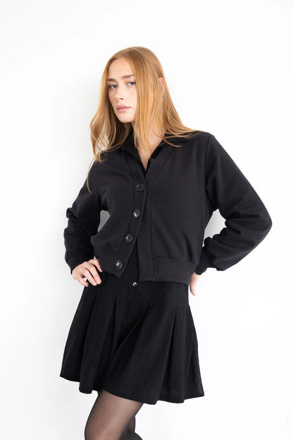 A woman wearing the Douglas Fleece Cardigan by Eve Lavoie in Black, a short cardigan with black buttons and a wide waistband. She is wearing it with a black skirt and standing against a white background
