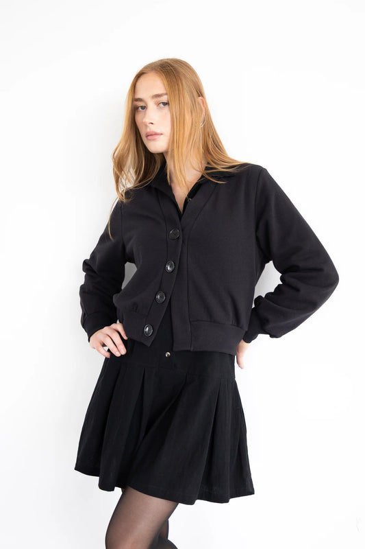 A woman wearing the Douglas Fleece Cardigan by Eve Lavoie in Black, a short cardigan with black buttons and a wide waistband. She is wearing it with a black skirt and standing against a white background