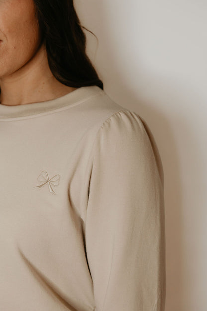 Close up of the monochromatic bow logo on the Tasia Sweatshirt by Betty x Bow in Crema, an oversized crew neck with subtly puffed shoulders and ribbing at the cuffs, hem and collar, 