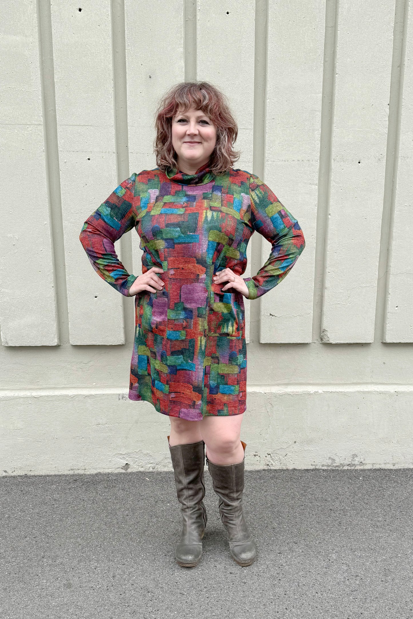 A  woman wearing the Drew Dress by Pure Essence in Multicolour Print, with a cowl neck, long sleeves, an above the knee hem, and patch pockets, standing outside in front of a white wall. 