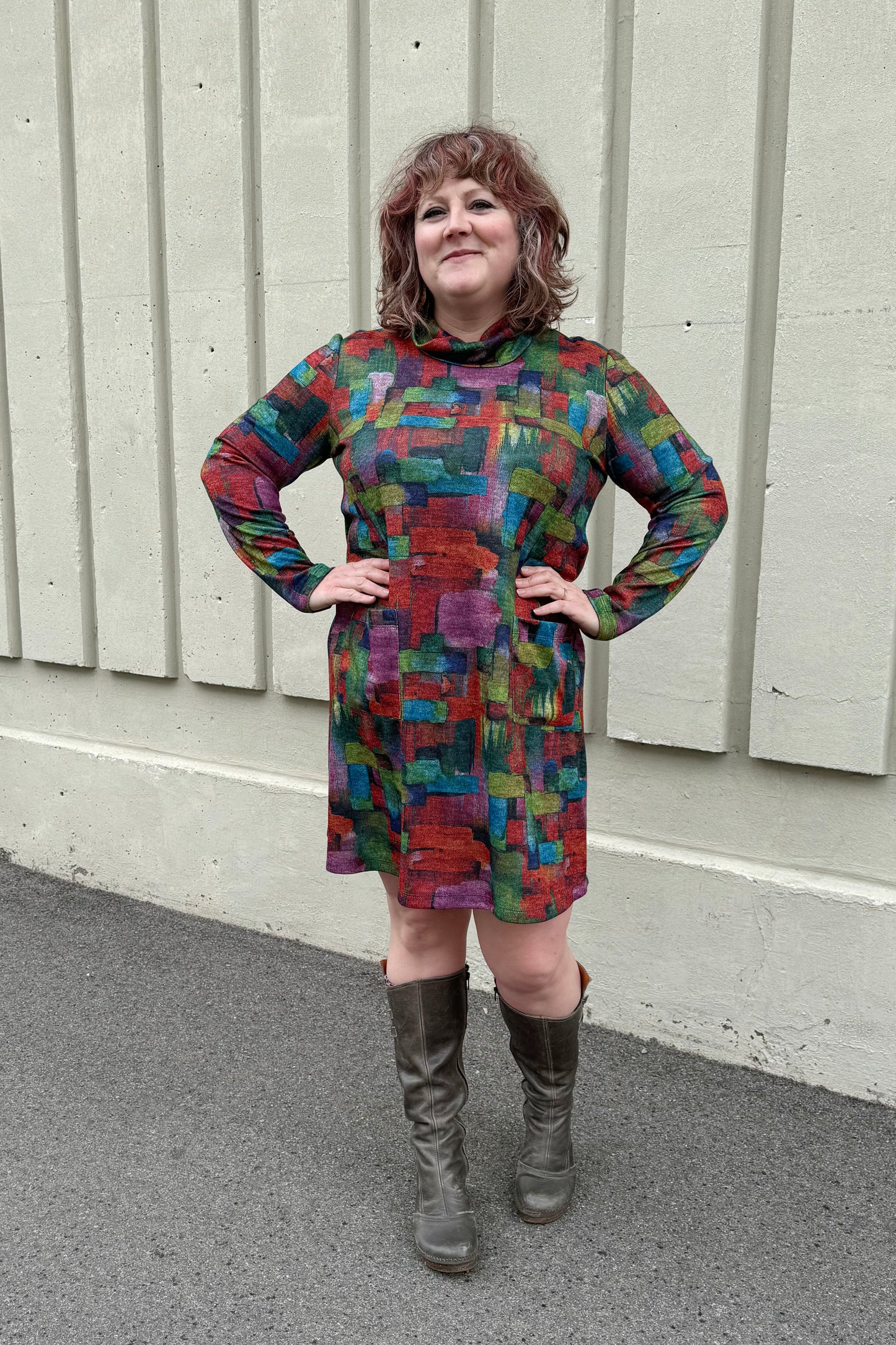 A  woman wearing the Drew Dress by Pure Essence in Multicolour Print, with a cowl neck, long sleeves, an above the knee hem, and patch pockets, standing outside in front of a white wall. 