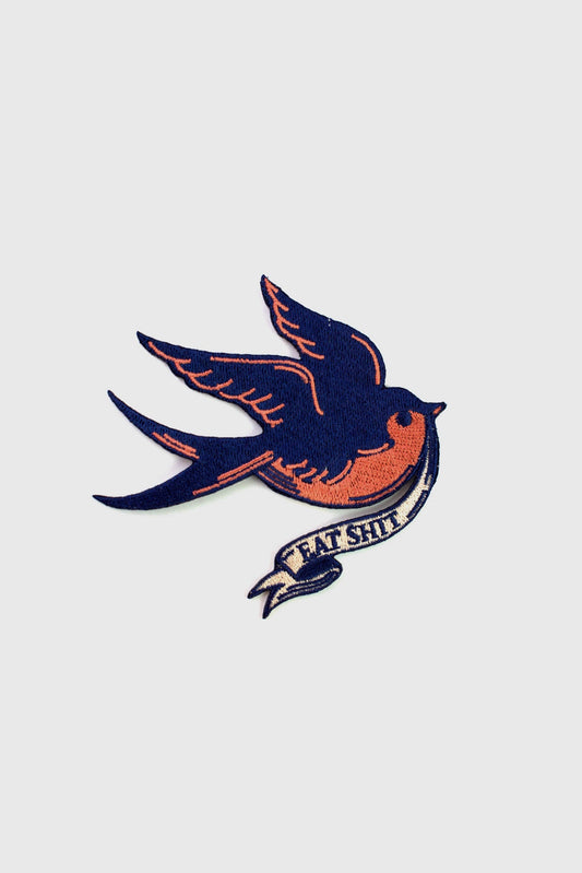 A vintage-style embroidered patch from Badaboöm Studio, 
featuring a flying bird in navy blue and orange, with a banner reading 'EAT SHIT' below, shown on a plain light grey background.