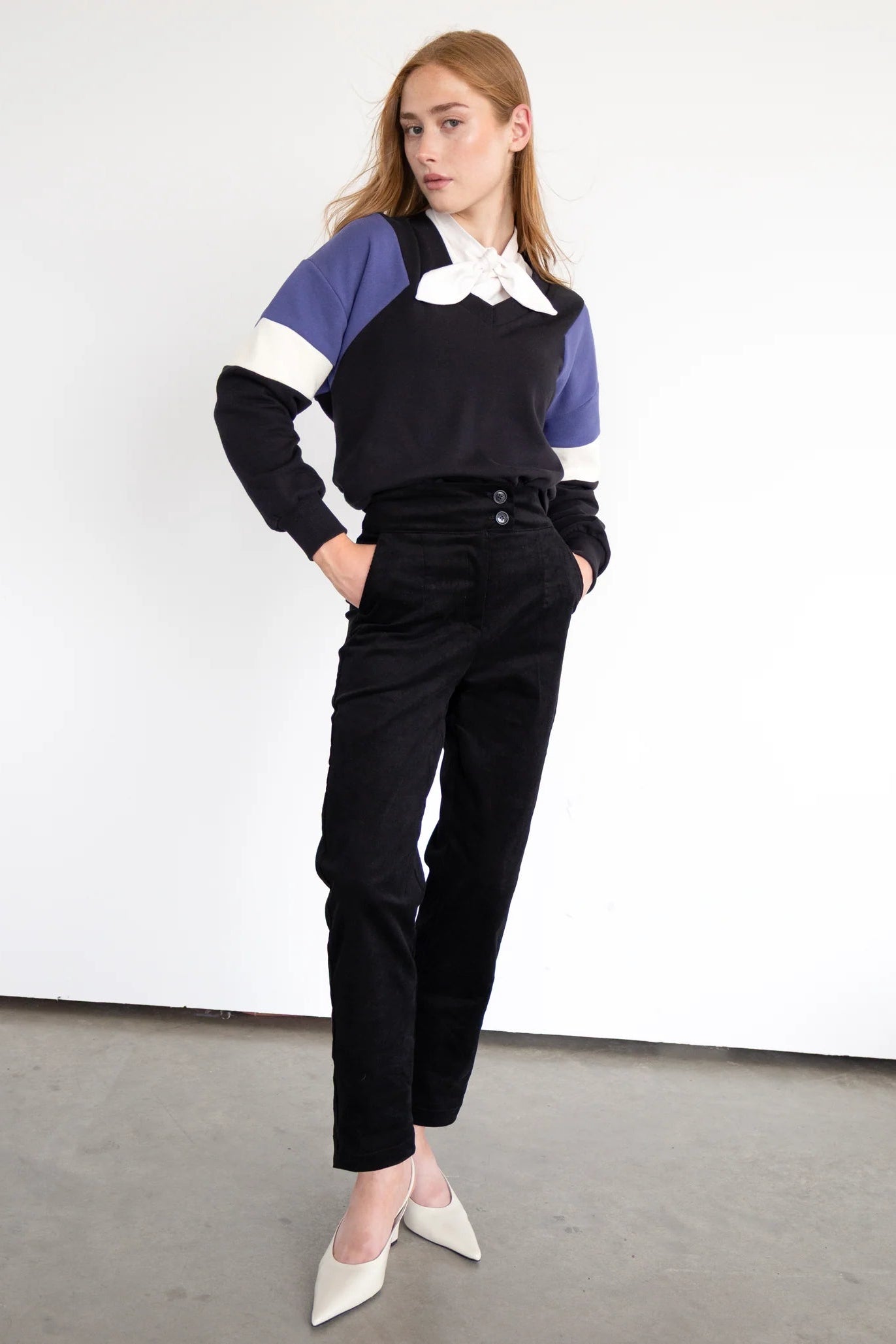 A woman wearing the Sinclair Sweatshirt by Eve Lavoie in Black, a relaxed sweatshirt with a V-neck and colour-blocked panels. She's wearing it with black pants and standing in front of a white background.