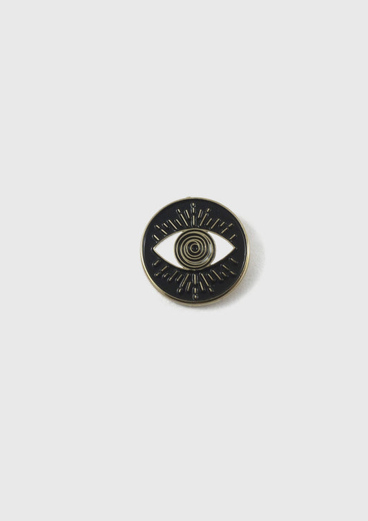 The Eyeball pin by Badaboöm Studio, a 
circular black enamel pin with a gold-tone metal border and an eye design in the centre, is shown against a white background.