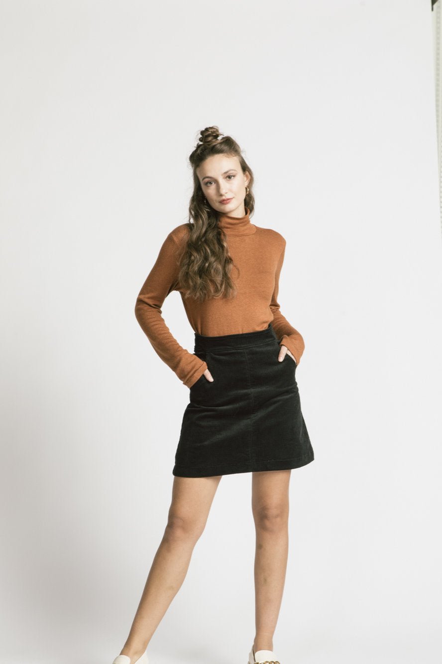 A woman wearing the Forever Turtleneck by Allison Wonderland in Caramel, with a black skirt, standing in front of a white background 