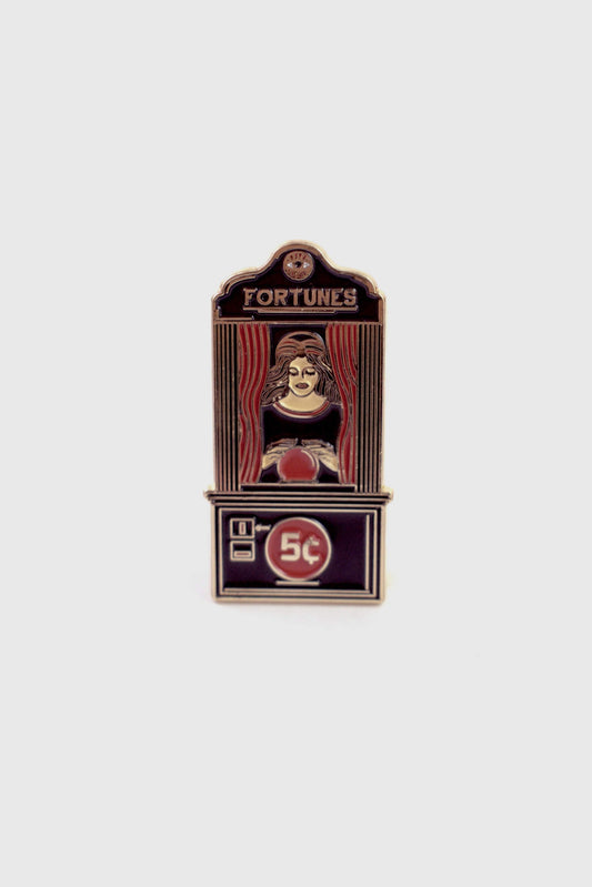 The Fortune Teller Pin from Badaboöm Studio is an enamel pin shaped like a vintage fortune telling booth with the word 'FORTUNES' at the top and '5c' on the lower panel. It's shown against a white background. 