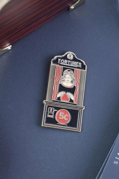The Fortune Teller Pin from Badaboöm Studio is an enamel pin shaped like a vintage fortune telling booth with the word 'FORTUNES' at the top and '5c' on the lower panel. It's shown against a navy background.