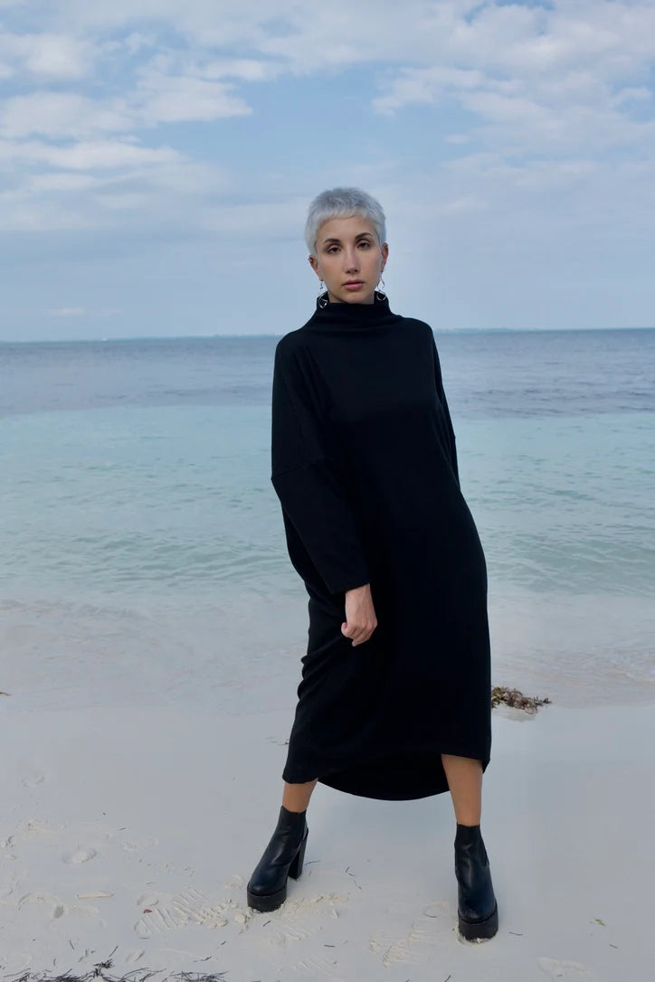Back view of a woman wearing the Frankfurt Dress by MAS in Pepper, a One Size ribbed cotton dress with a mock turtleneck, long batwing sleeves, and midi length. She is standing on a beach.