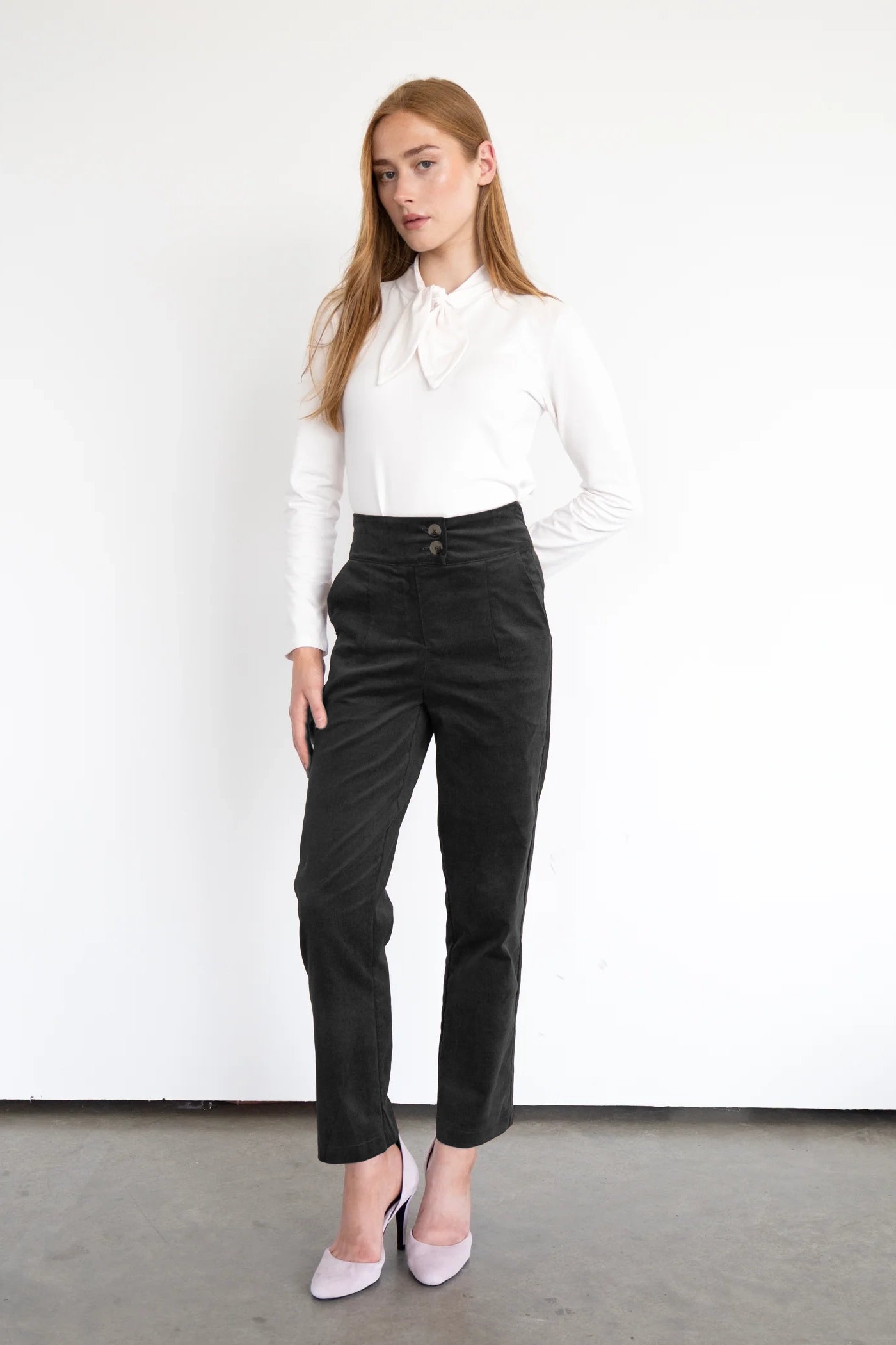 A woman wearing the Graham Corduroy Pants by Eve Lavoie in Black, with a high waist, double button, slim fit and ankle length leg. She is wearing them with a white top and standing in front of a white background. 