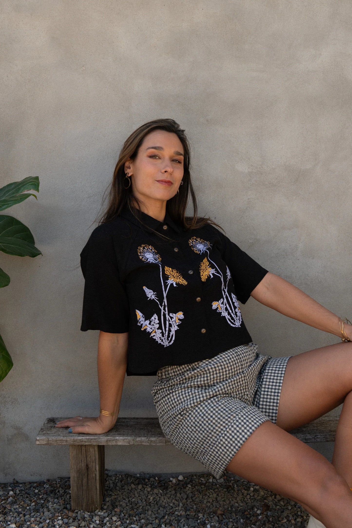 A woman wearing the Hosta Screen-printed Blouse by Kazak in Black with Dandelions, with hand-screen-printed flowers, mid-length raglan sleeves, a classic collar, and a button front. She is wearing it with gingham shorts and sitting outside by a grey wall. 