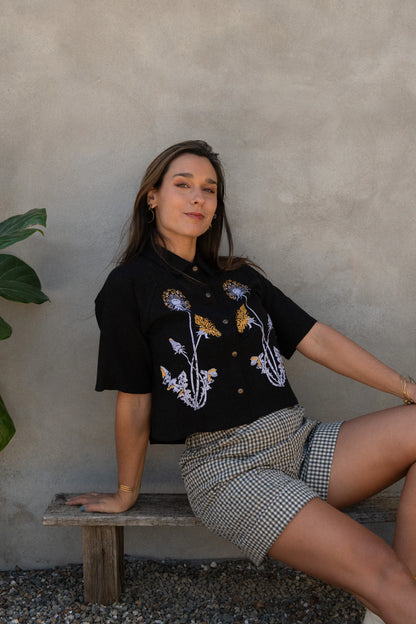 A woman wearing the Hosta Screen-printed Blouse by Kazak in Black with Dandelions, with hand-screen-printed flowers, mid-length raglan sleeves, a classic collar, and a button front. She is wearing it with gingham shorts and sitting outside by a grey wall. 