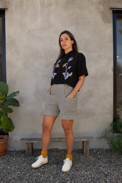 A woman wearing the Hosta Screen-printed Blouse by Kazak in Black with Dandelions, with hand-screen-printed flowers, mid-length raglan sleeves, a classic collar, and a button front. She is wearing it with gingham shorts and standing outside by a grey wall. 