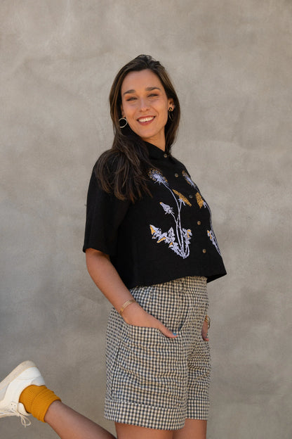A woman wearing the Hosta Screen-printed Blouse by Kazak in Black with Dandelions, with hand-screen-printed flowers, mid-length raglan sleeves, a classic collar, and a button front. She is wearing it with gingham shorts and standing outside by a grey wall. 