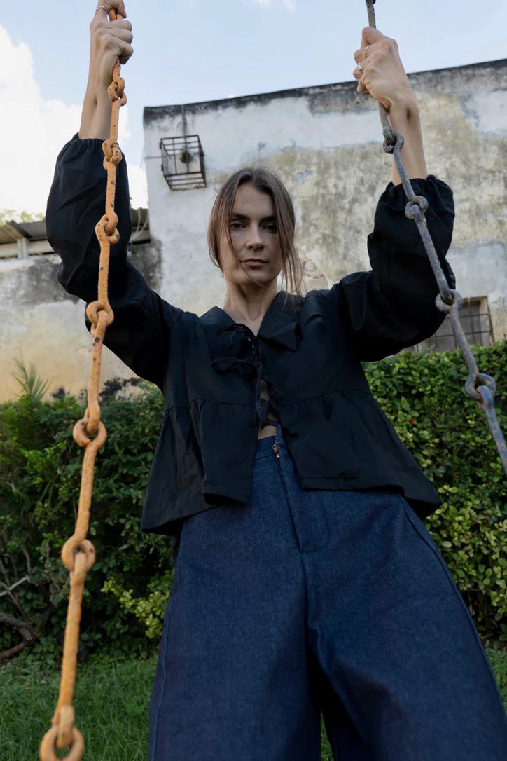 A woman wearing the Vienna Pants in Jeans by MAS, high waisted pants with wide legs and a zipper and tab closure. She is wearing them with a black top and sitting on a swing.