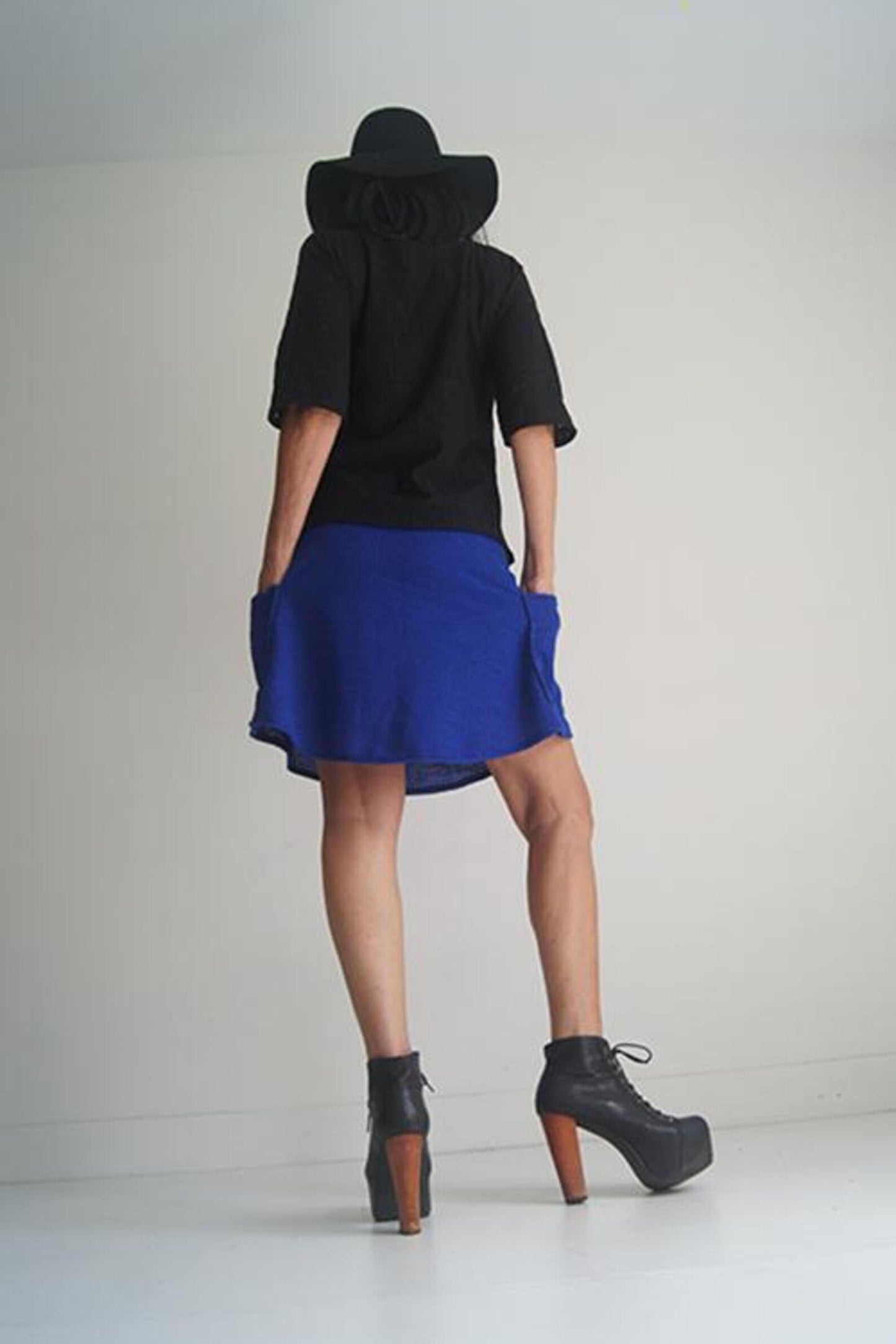 Back view of a woman wearing the Short Skirt by SI Design in Blue, an above-the-knee A-line skirt with an elastic waist and patch pockets. She is wearing it with a black top and standing in front of a grey background. 