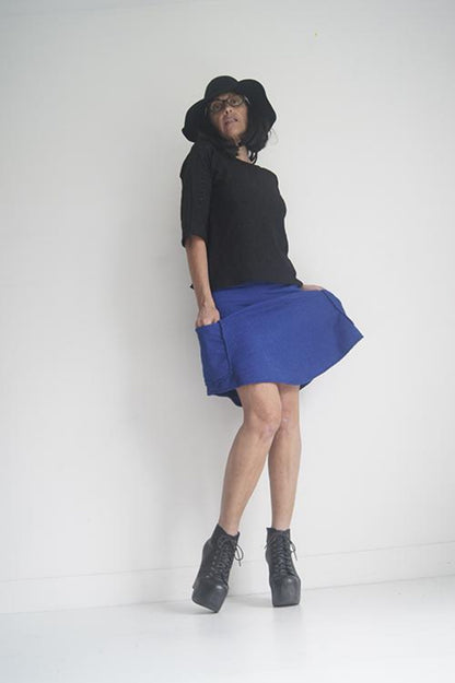 A woman wearing the Short Skirt by SI Design in Blue, an above-the-knee A-line skirt with an elastic waist and patch pockets. She is wearing it with a black top and standing in front of a grey background. 