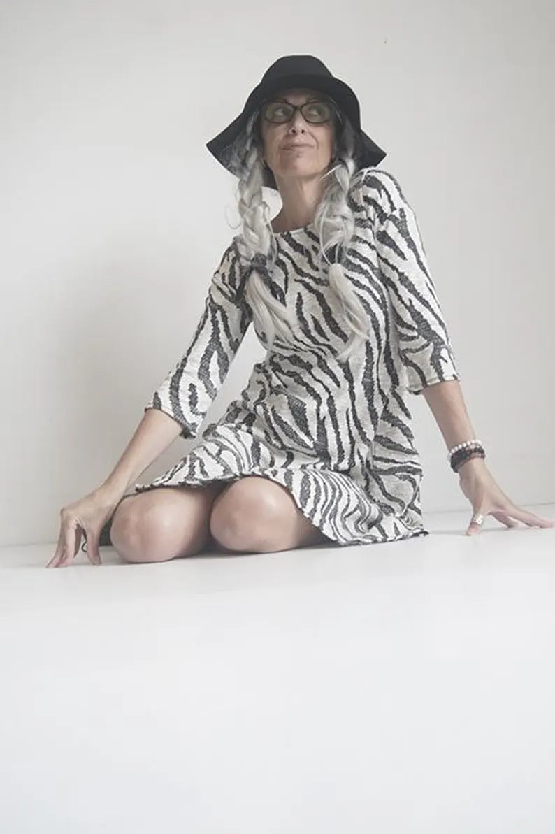 A woman wearing the Casual Dress by SI Design in Ivory/Black, with a round neck, 3/4 sleeves, a loose fitting A-line shape, and a knee length, sitting in front of a white background 