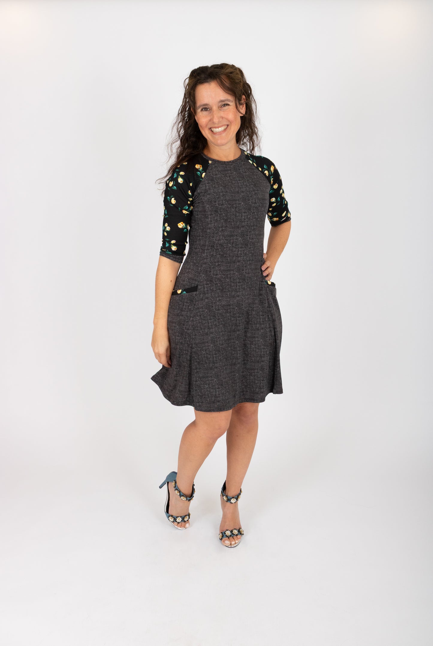 A woman wearing the Raphaelle Dress from Infime in Black, an A-line knee-length dress with 3/4 raglan sleeves that feature a floral print and pockets with a floral detail. She is standing in front of a white background