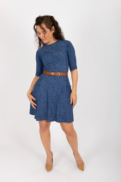 A woman wearing the Raphaelle Dress from Infime in Blue, an A-line knee-length dress with 3/4 raglan sleeves and pockets. She is standing in front of a white background.