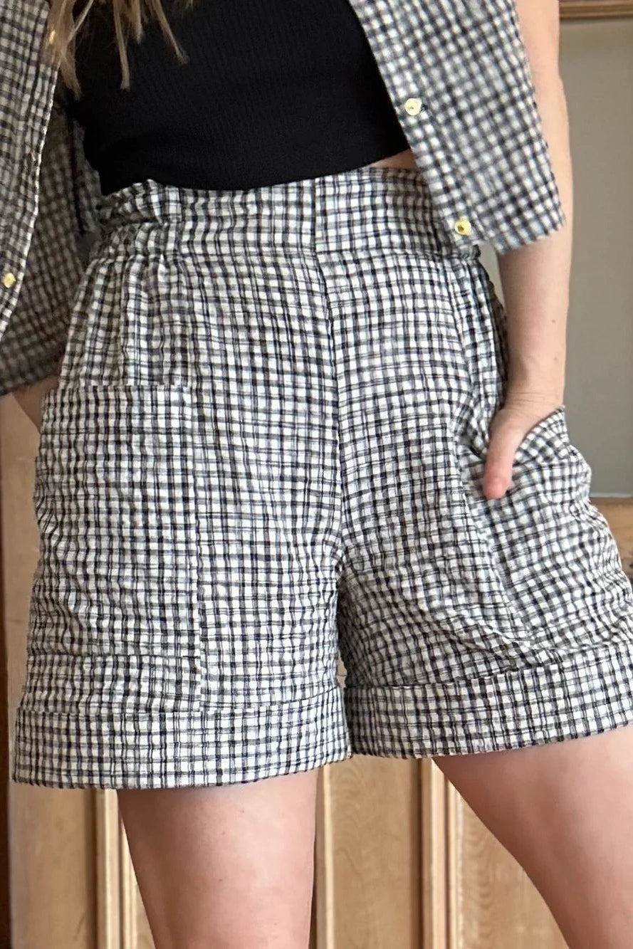 Close up view of a woman wearing the Iris Shorts by Kazak in Black and Cream Vichy/Gingham, with a smooth front waist, elastic at the back, large pockets, and mid-thigh length. She is wearing them with a black tank and matching open top and standing in a room with a door. 