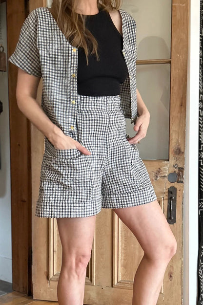 Neck down view of a woman wearing the Iris Shorts by Kazak in Black and Cream Vichy/Gingham, with a smooth front waist, elastic at the back, large pockets, and mid-thigh length. She is wearing them with a black tank and matching open top and standing in a room with a door. 