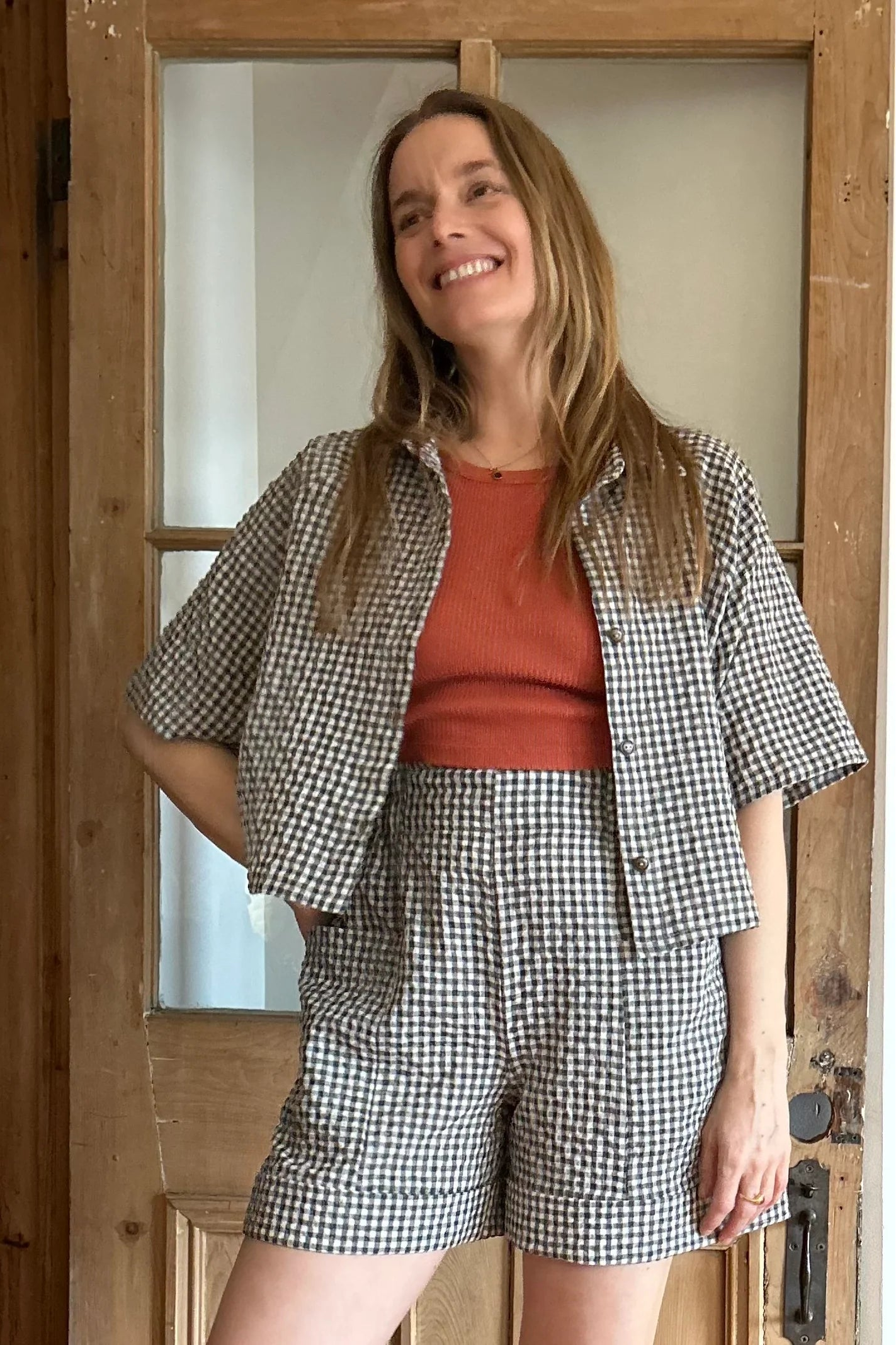 A woman wearing the Iris Shorts by Kazak in Teal Vichy/Gingham, with a smooth front waist, elastic at the back, large pockets, and mid-thigh length. She is wearing them with an orange tank and matching open top and standing in a room with a door. 