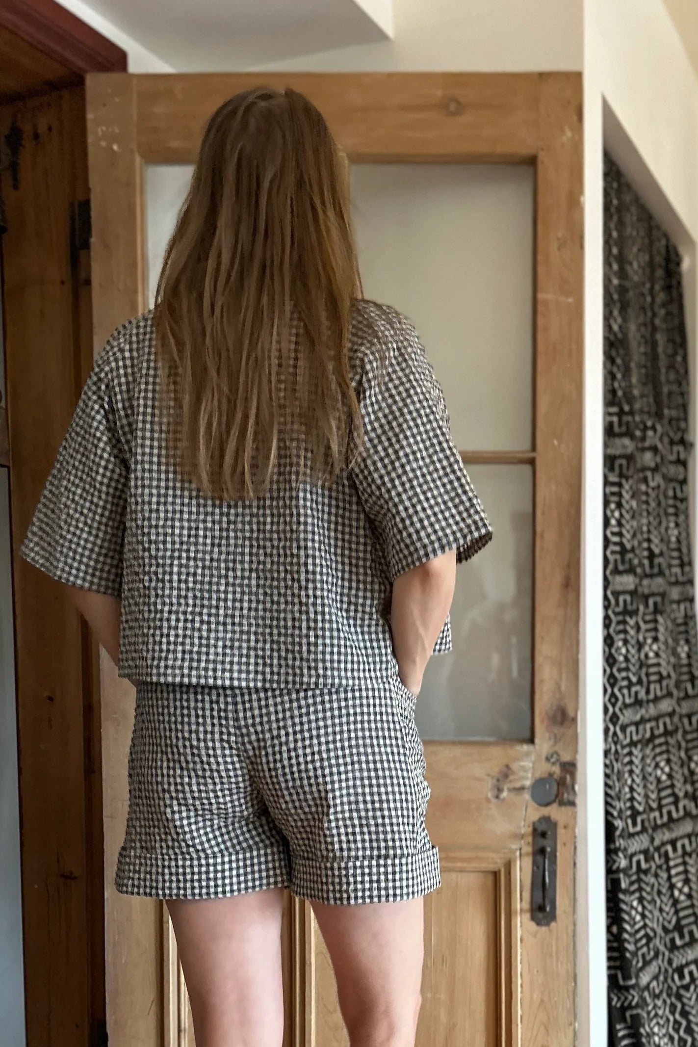 Back view of a woman wearing the Iris Shorts by Kazak in Teal Vichy/Gingham, with a smooth front waist, elastic at the back, large pockets, and mid-thigh length. She is wearing them with an orange tank and matching open top and standing in a room with a door. 
