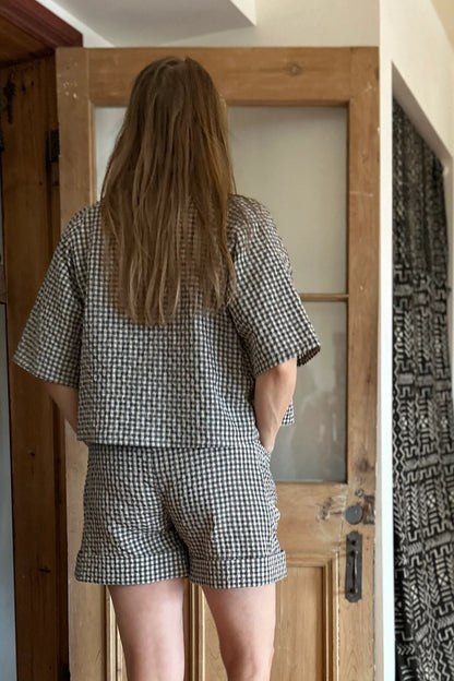 Back view of a woman wearing the Iris Shorts by Kazak in Teal Vichy/Gingham, with a smooth front waist, elastic at the back, large pockets, and mid-thigh length. She is wearing them with an orange tank and matching open top and standing in a room with a door. 